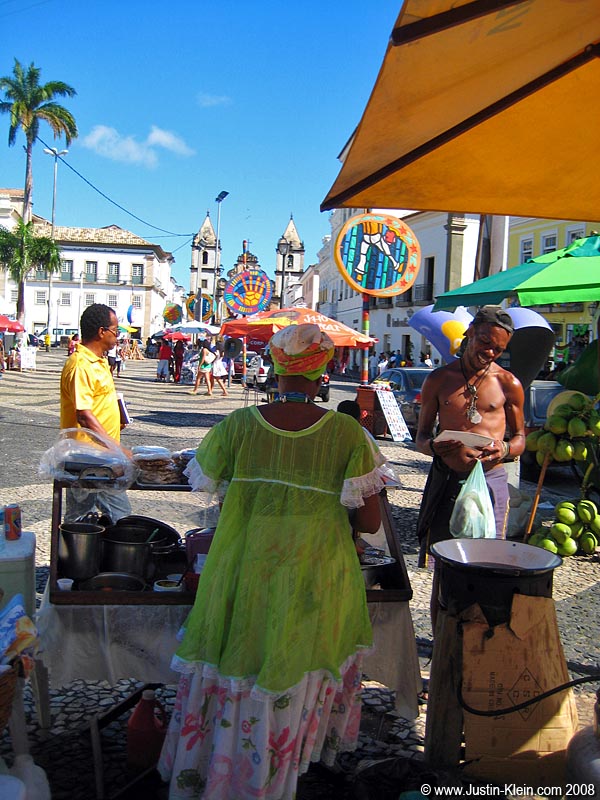 A man buys a light snack from a woman in traditional Bahian attire.